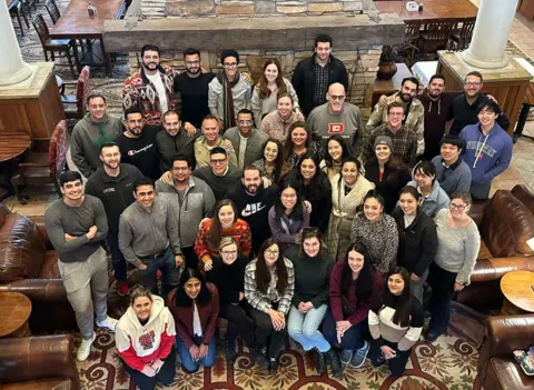 A large group of people smiling and posing together in a high-angle group photo inside a rustic lodge lobby with leather armchairs and a stone fireplace.