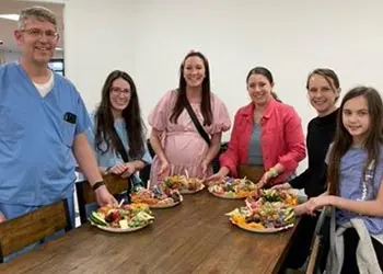 A group of six people standing around a wooden dining table displaying decorative charcuterie plates.
