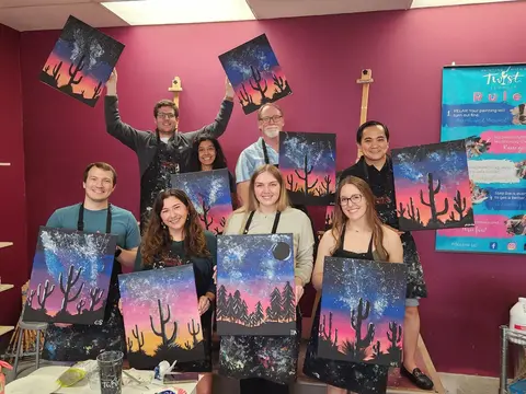 A group of eight people wearing aprons holding up their finished paintings of desert landscapes at a Painting with a Twist art studio.