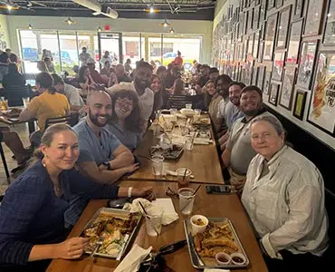 A group of medical residents sitting along a long table inside a busy restaurant, smiling for a group photo.