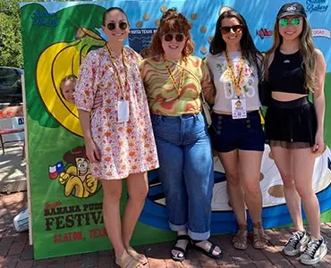Four women posing together outdoors in front of a Slaton Banana Pudding Festival photo backdrop.