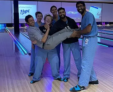 A group of medical residents in blue scrubs laughing and posing together on a bowling lane, holding one colleague in their arms.