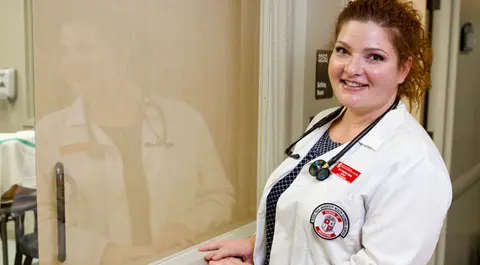 Medical student wearing a white coat and stethoscope in a clinical setting.