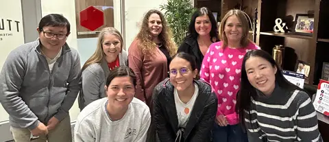 Eight TTUHSC employees smile for a group photo inside an office lobby. The group is gathered in front of a bookshelf with framed photos and décor, with several standing and others leaning toward the camera. One person wears a pink sweater with white hearts, while the others wear casual sweaters, jackets, and business-casual attire.