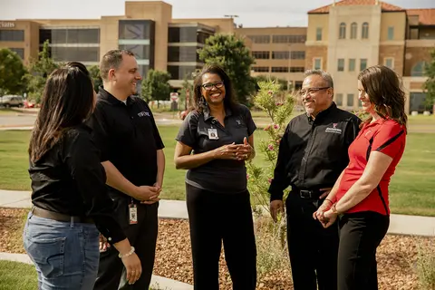 Five TTUHSC employees stand in a circle outdoors on campus, smiling and talking together on a grassy lawn with academic buildings in the background.