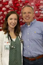 A female medical student wearing a white lab coat and stethoscope smiling alongside an alumnus in front of a red decorative backdrop.
