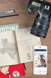Top-down flat lay view of a wooden desk featuring a DSLR camera, an open sketchbook with hand-drawn lettering, markers, printed magazines, and a smartphone displaying a social media feed.