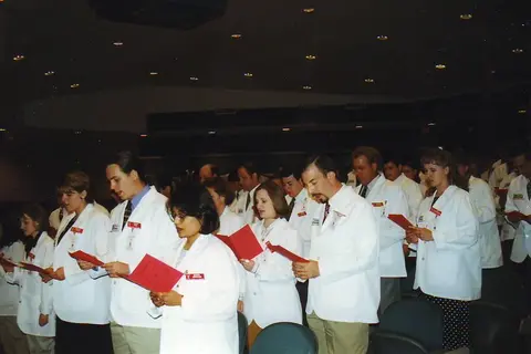 Pharmacy students in white coats recite an oath while holding red booklets during a ceremony.