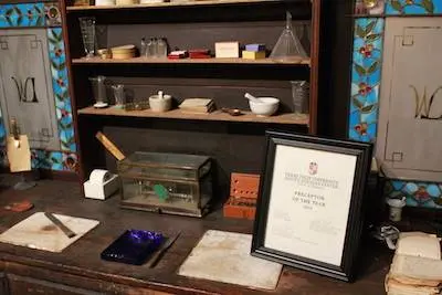 Historic pharmacy workbench displaying glassware, a mortar and pestle, tools and a framed prescription.
