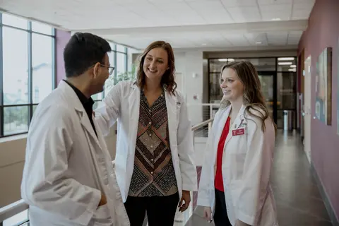 Three pharmacy professionals in white coats talk in a bright university hallway.