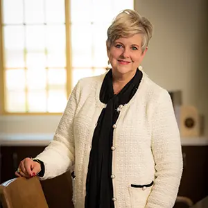 Portrait of a TTUHSC administrator standing beside a chair in a conference room, wearing a white textured blazer with black trim and a black scarf, smiling toward the camera.