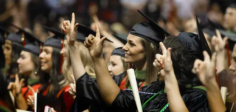 Smiling Texas Tech University Health Sciences Center graduates in graduation gowns, black caps, and honor cords throwing the Texas Tech "Guns Up" hand sign while holding diplomas in a crowded auditorium.
