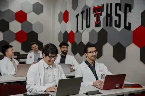 Medical students in white coats work on laptops in a TTUHSC classroom.