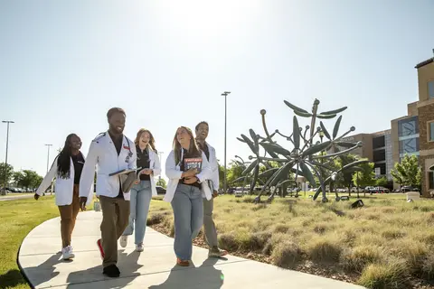Five TTUHSC students in white coats walking together near an outdoor sculpture on campus.