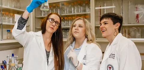 Three researchers in lab coats examine a flask together in a laboratory.