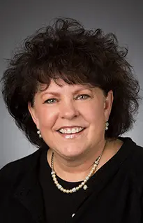 Headshot of a smiling Rebeca Geist with dark curly hair wearing a black top and a pearl necklace.