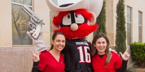 Texas Tech’s Raider Red mascot poses with two students in red scrubs outside a campus building.