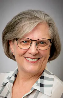 Headshot of a smiling Gini Holter with short gray hair and glasses, wearing a patterned collared shirt.