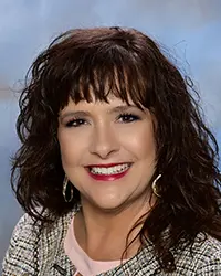 Headshot of Jodi Fry with dark wavy hair and red lipstick, wearing a patterned jacket and gold hoop earrings.