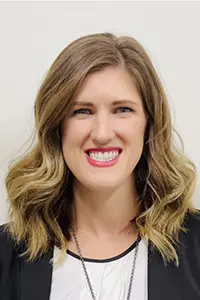 Headshot of a smiling woman with shoulder-length wavy light brown hair, wearing a white top and black jacket.