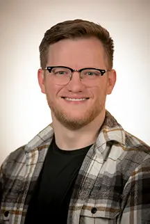 Headshot of Logan Gaines wearing glasses and an unbuttoned plaid shirt over a black t-shirt, smiling at the camera.