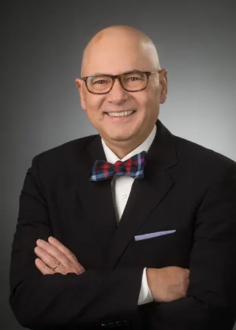 Professional portrait of a the som wearing glasses, a dark suit and a plaid bow tie.