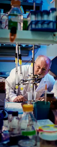 Researcher in a laboratory examining equipment surrounded by bottles and scientific instruments.