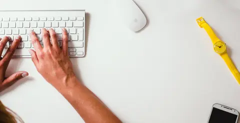 Hands typing on a white keyboard beside a mouse, yellow watch and smartphone.