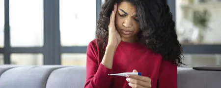 A young woman with long, curly dark hair wearing a red shirt holds a digital thermometer, looking down at it with a worried expression while holding one hand to her temple.