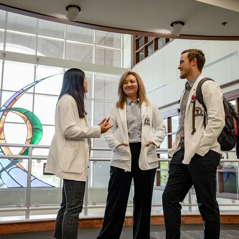 TTUHSC Amarillo medical students wearing white coats standing in front of windows talking and smiling.