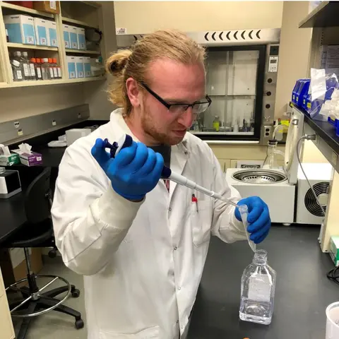 A scientist wearing a white lab coat and blue protective gloves uses a pipette to transfer liquid into a small laboratory tube. He is working at a laboratory bench surrounded by scientific equipment, bottles, and storage containers.