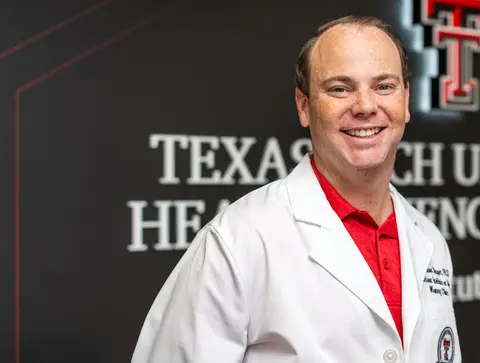 Jonathan Singer wears a white coat and stands in front of a wall decorated with the TTUHSC logo.