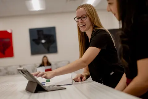 Smiling woman in glasses reaches across a conference table to use a tablet beside an open laptop and open textbook, while colleagues watch in a bright office.