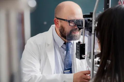 A bearded eye-care professional in a white lab coat examines a patient through an optical instrument, with the patient partially visible in the foreground.