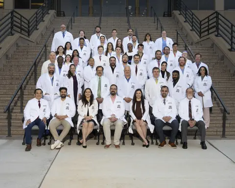 A large group of medical residents, faculty, and clinical staff posing together outdoors on the wide stone steps of a building entrance. The group consists of men and women of diverse backgrounds, dressed in white lab coats over professional attire or medical scrubs. The front row is seated in office chairs, while the remaining members stand arranged in tiers up the staircase, framing the central group against the building facade.