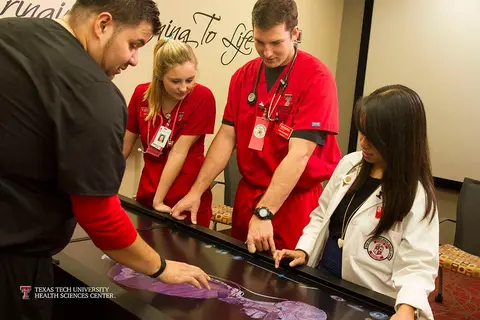 Four healthcare students in red and white medical uniforms gather around a table, examining a large anatomical image under a transparent surface in a classroom or training setting.