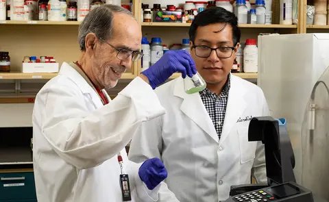 Two scientists in white lab coats work together in a laboratory. The older scientist on the left holds up a glass flask containing a green liquid while wearing purple gloves, and a younger scientist on the right looks on. Shelves filled with laboratory bottles and containers are visible in the background, along with scientific equipment in the foreground.