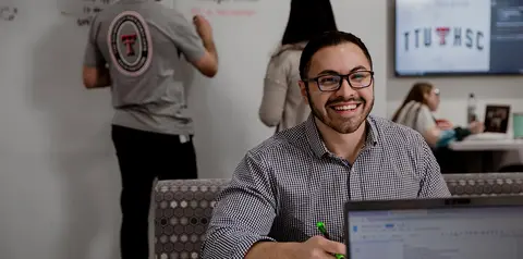 A smiling man wearing glasses and a checkered shirt sits at a laptop in an office, holding a green pen. In the background, coworkers work at a whiteboard and desks.