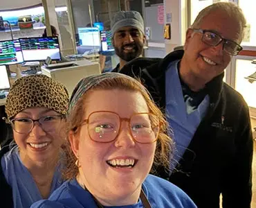 Four smiling healthcare workers pose for a selfie in a hospital or clinical setting, with medical equipment and computer monitors visible in the background.