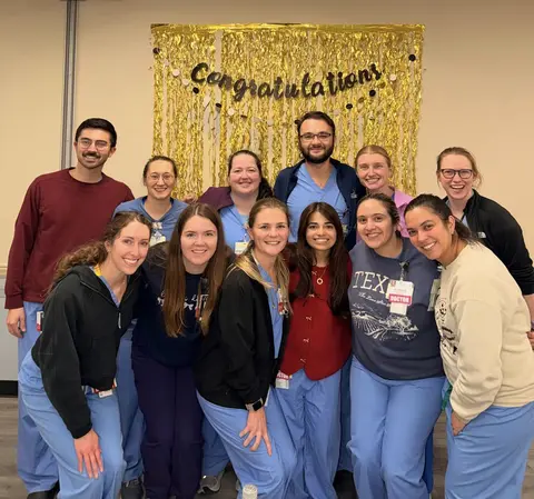 A cheerful group of twelve healthcare professionals, mostly dressed in light blue scrubs with various jackets and layered long-sleeve shirts, posing together for a celebratory photo. They are gathered in front of a shiny gold fringe backdrop adorned with a black "Congratulations" banner.