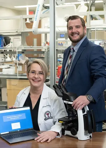 one person sitting, one standing, posing with a laptop in a lab