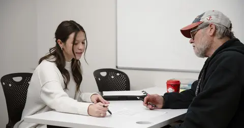 Woman sitting across from a man, indicating something on the table between them with a pen