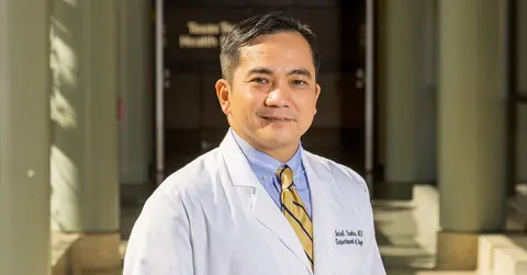 Male surgeon wears a white coat and stands in a hallway.