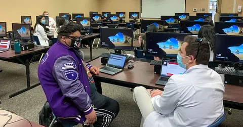 high school student sitting with mentor in computer lab, both in masks