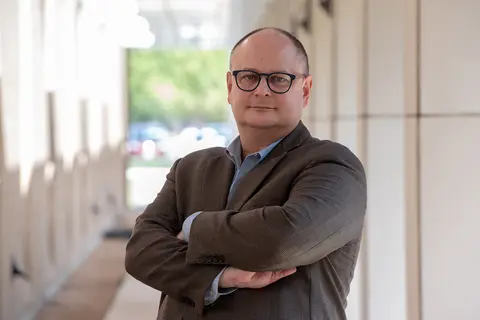 Man wears a brown sports coat and stands with his arms crossed.