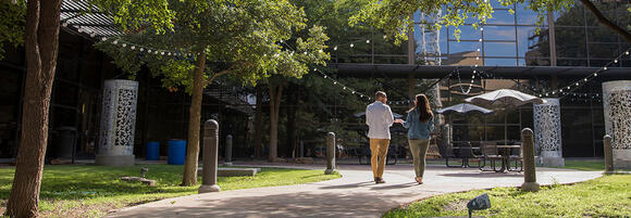Two people walk along a courtyard path outside a modern TTUHSC building, with trees, patio seating and string lights overhead.